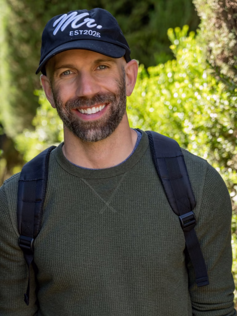 Man with beard wearing a "Mr. EST2025" hat and backpack, smiling.