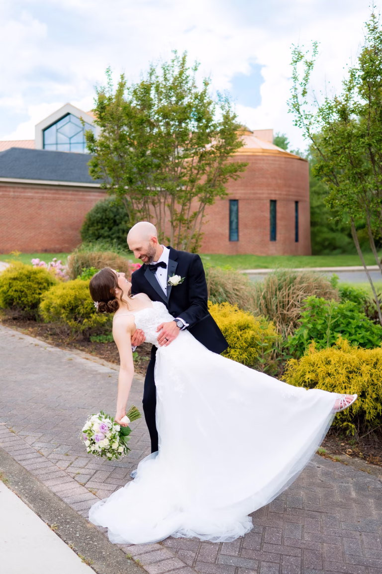 Bride in white dress dips back as groom smiles at her.