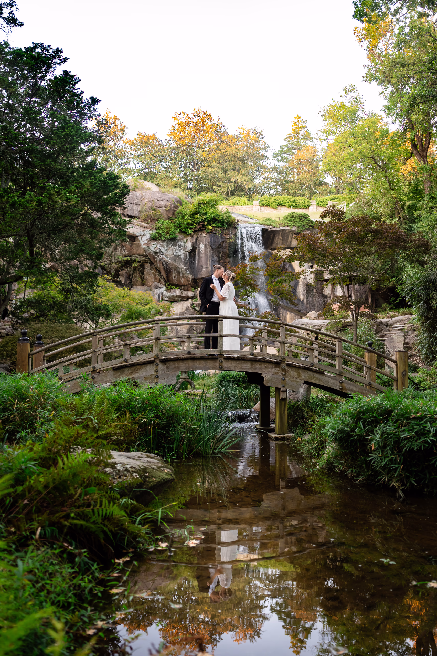 Bride and groom kiss on wooden bridge with waterfall backdrop.
