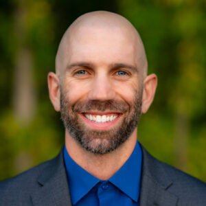 Smiling man with beard in blue shirt and grey suit jacket against green background.