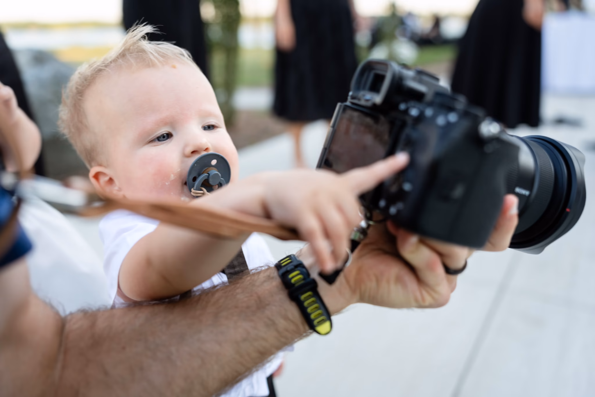 Baby with pacifier reaches for a DSLR camera held by an adult.