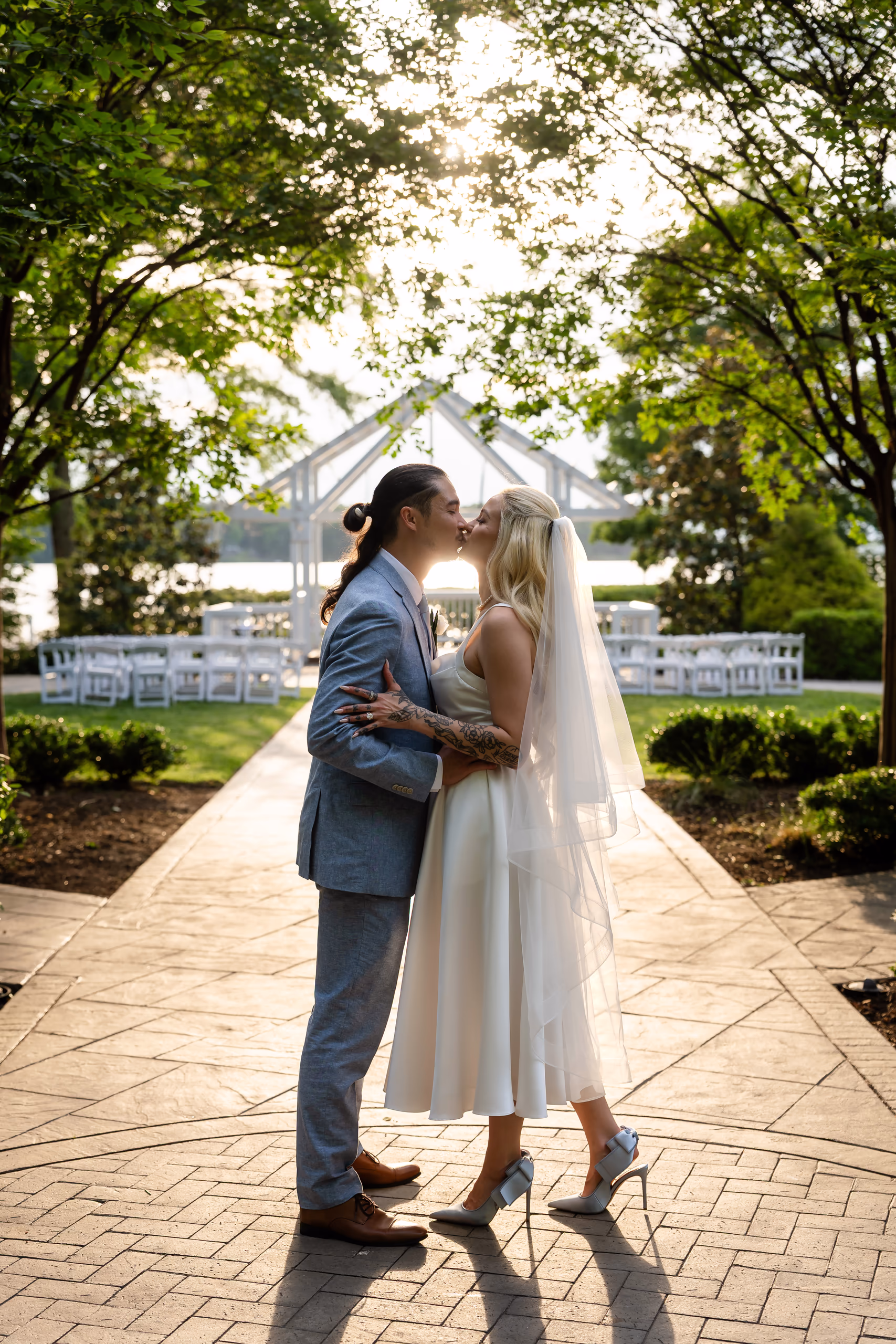 Bride and groom kissing at sunset on a paved pathway with trees and wedding ceremony setup