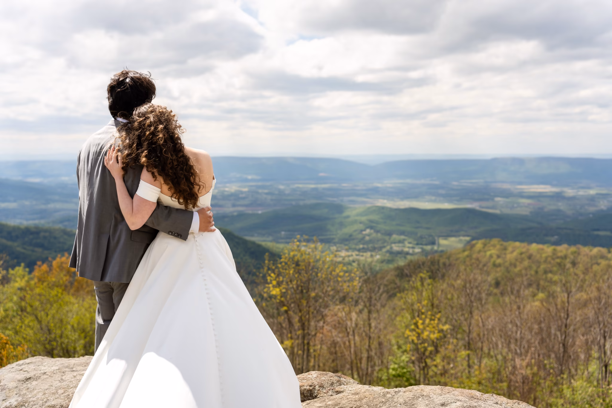 Bride and groom embrace on a mountain overlook, gazing at a scenic valley below.