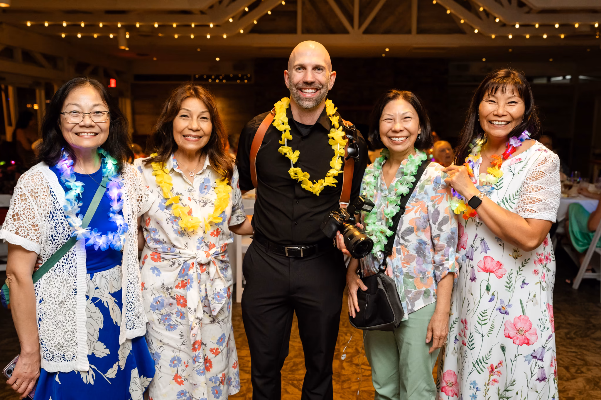 Bald man with leis and camera poses with four smiling women in floral dresses.