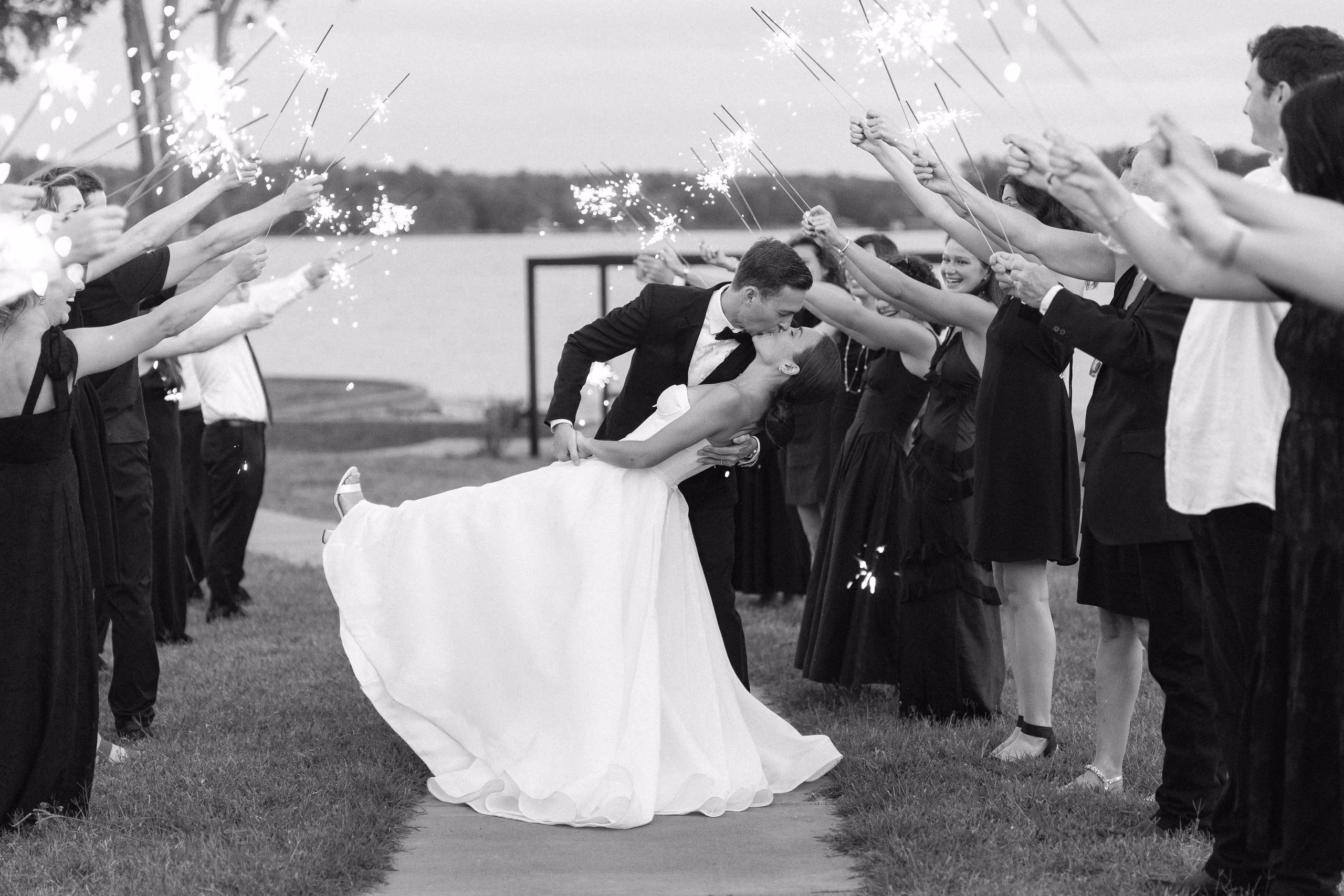 Bride and groom dip and kiss surrounded by guests holding sparklers at dusk.